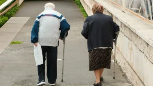 A poignant shot capturing an elderly couple walking away from the camera along a paved, inclined path. Both individuals rely on crutches for mobility, symbolizing mutual support and resilience in old age. The man, in a light tracksuit top, holds a small bag, while the woman wears a dark blazer and a patterned skirt. This image is suitable for concepts related to aging, healthcare, companionship, physical therapy, senior lifestyle, and the journey of life. elderly, senior, couple, walking, crutches, mobility, aging, health, healthcare, support, companionship, old age, lifestyle, path, outdoor