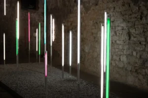 Vertical fluorescent lights in various colors illuminate a stone wall.