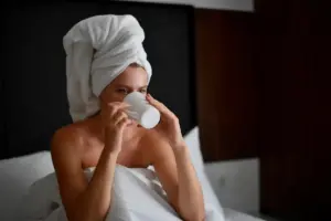 Young blonde woman with a white towel wrapped around her head drinking hot coffee in bed during her morning routine.